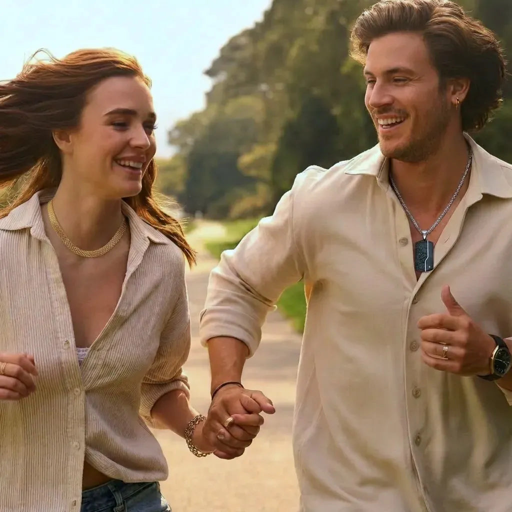Couple heureux main dans la main courant sur un chemin ensoleillé. Chemises beiges, collier pendentif homme, collier chaîne doré femme. Joie et bijoux minimalistes.