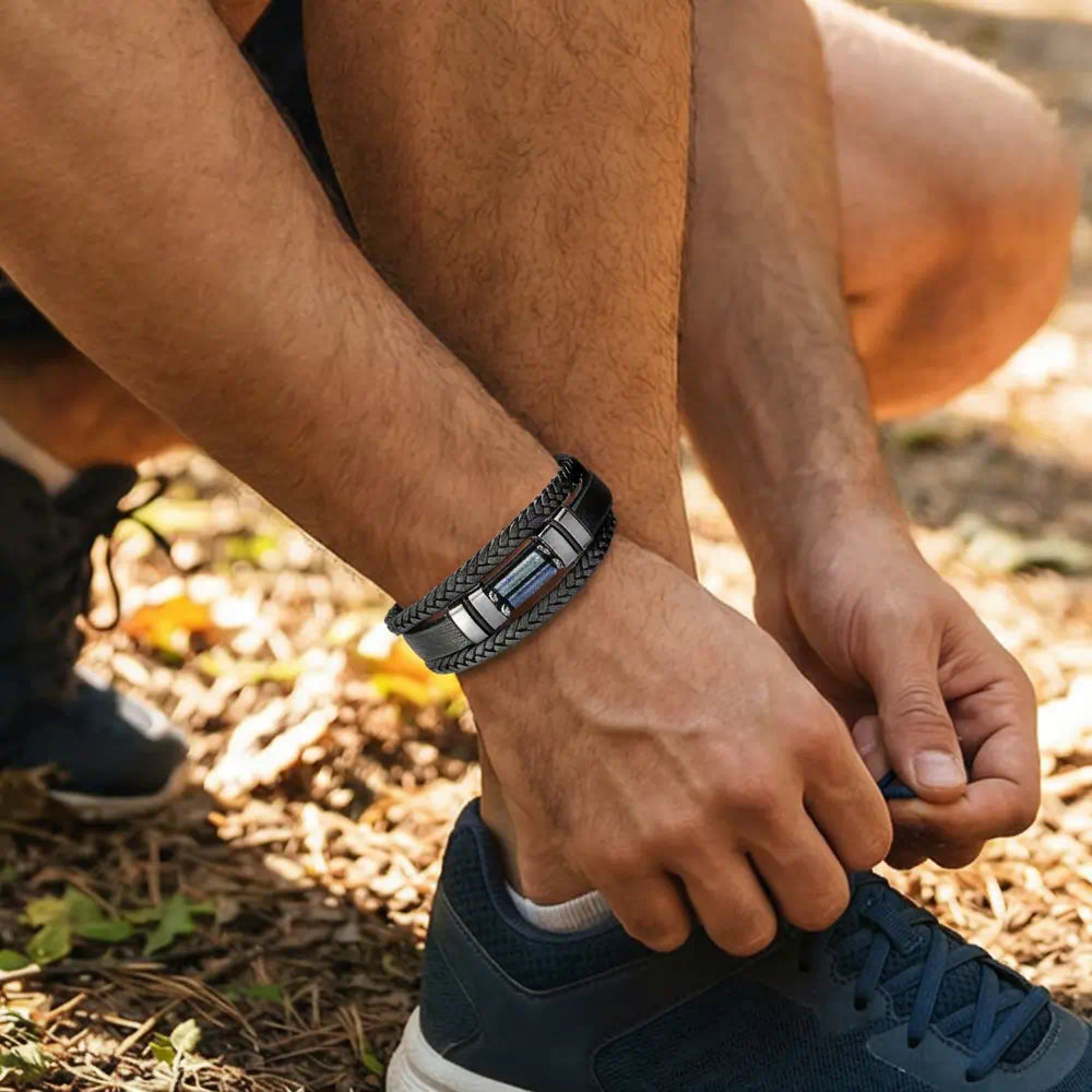 Zoom immersif sur le bracelet Azimut au poignet d'un sportif ajustant ses lacets ; les lanières de cuir noir et les pierres bleues contrastent avec l'éclat solaire du sentier forestier en été.