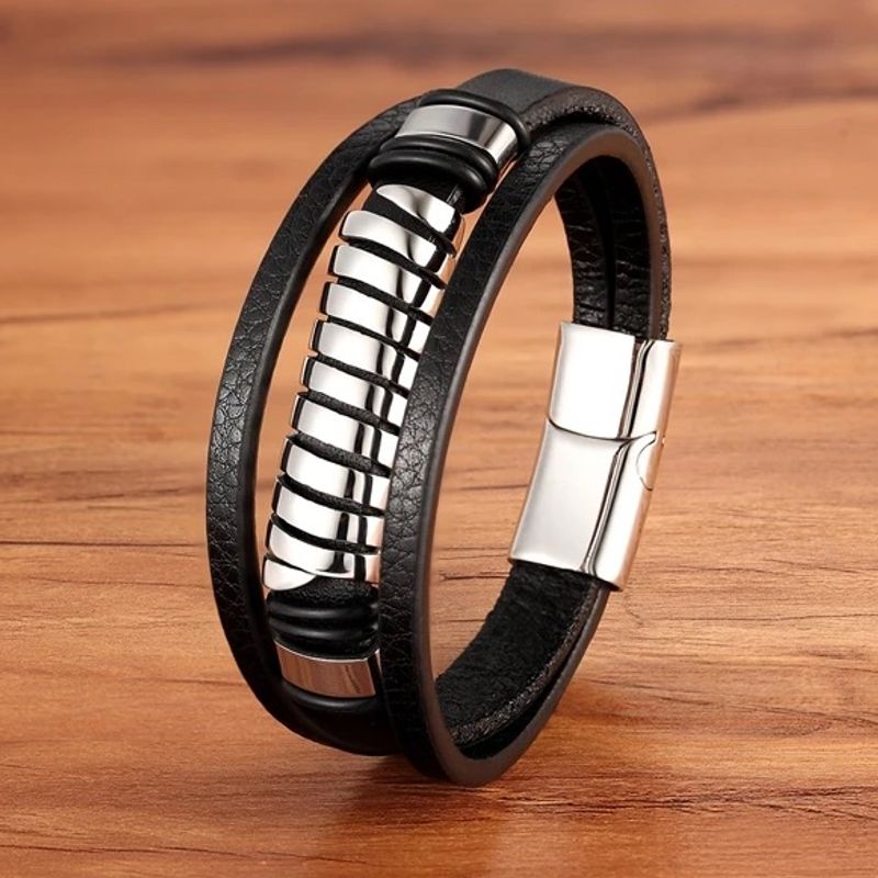 Bracelet spirale acier avec perle tube ajourée, argentée et multilanère en cuir noir. Fermoir magnétique en acier inoxydable de couleur argent. Ce wrap est présenté fermé, dressé sur un plan de travail en bois clair.