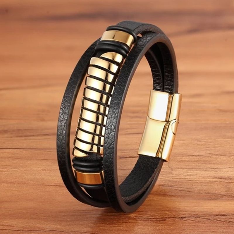 Bracelet spirale acier avec perle tube ajourée, vis sans fin, dorée et multilanère en cuir noir. Fermoir magnétique en acier inoxydable de couleur or. Ce wrap est présenté fermé, dressé sur un plan de travail en bois clair.