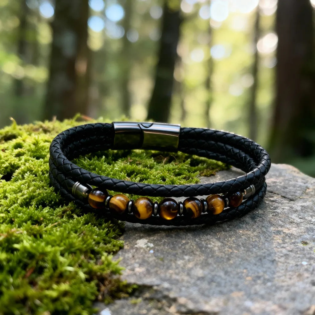 Plan large du bracelet Talis posé à plat, symbolisant l'ancrage. Le multi-rangs en cuir souple noir et l'œil de tigre reposent sur la mousse et le rocher, exprimant la force naturelle.