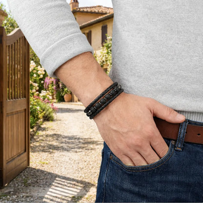 Focus détaillé sur le bracelet Vulcan en cuir noir tressé et perles de pierre de lave, porté élégamment avec un pull gris sur fond de propriété résidentielle.