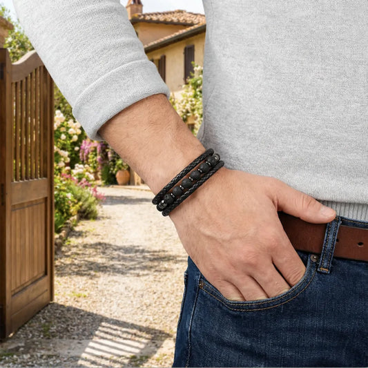 Focus détaillé sur le bracelet Vulcan en cuir noir tressé et perles de pierre de lave, porté élégamment avec un pull gris sur fond de propriété résidentielle.